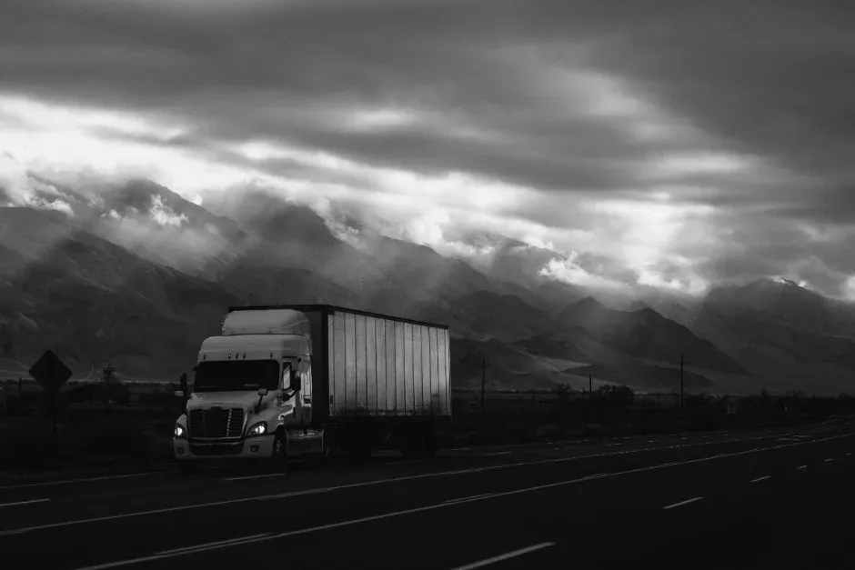 Semi truck hauling a trailer on a highway beneath cloud-covered mountains
